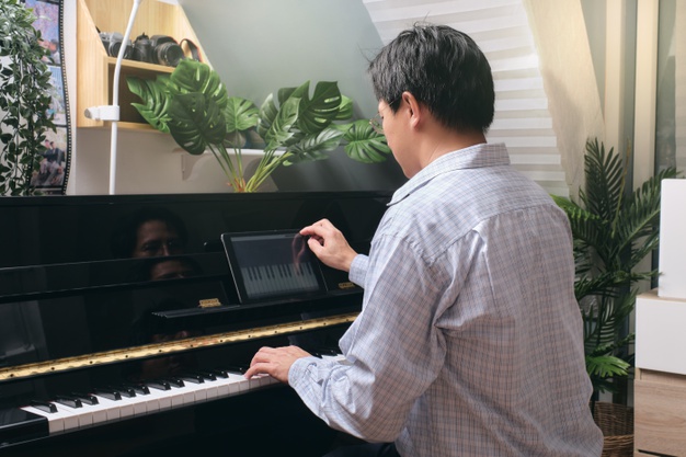 hombre-adulto-aprendiendo-tocar-piano-usando-tableta-digital-leccion-linea-curso-sala-estar-casa-feliz-empresario-asiatico-relajandose-tocando-piano_83369-411